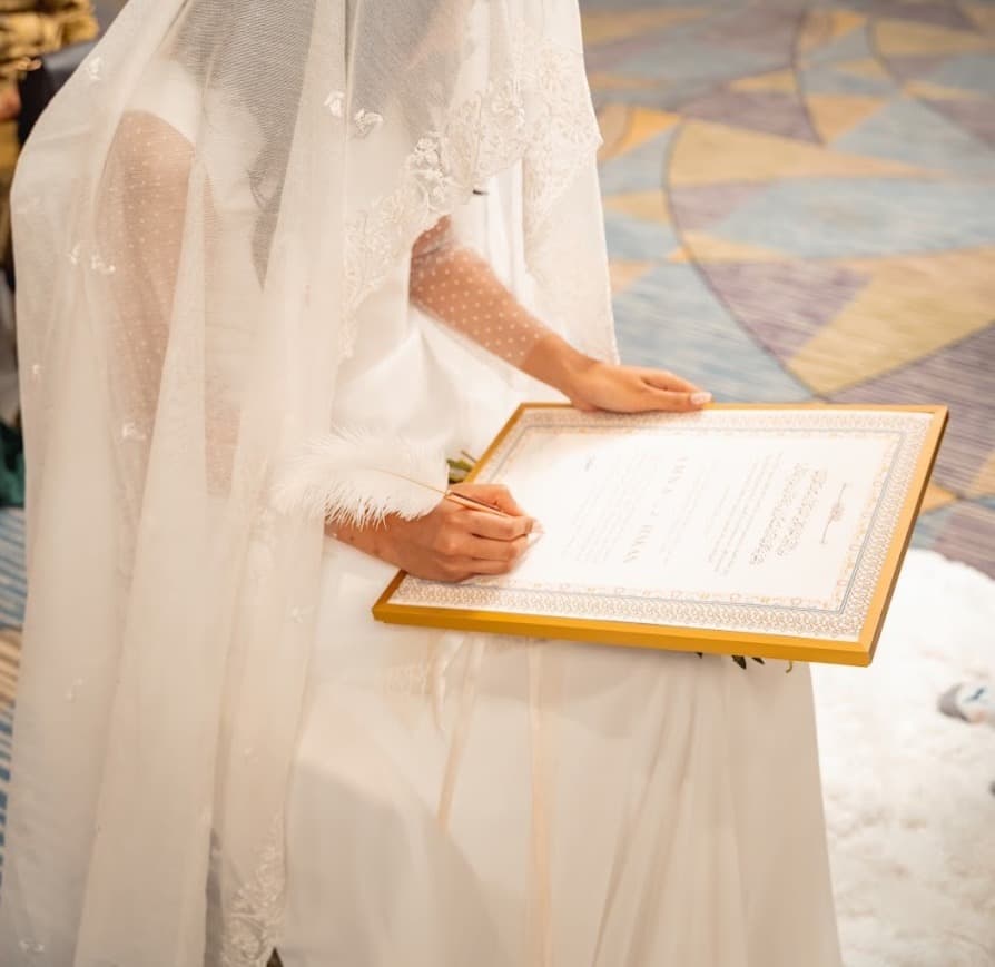 A bride in a white gown signing her large gold-framed nikah certificate