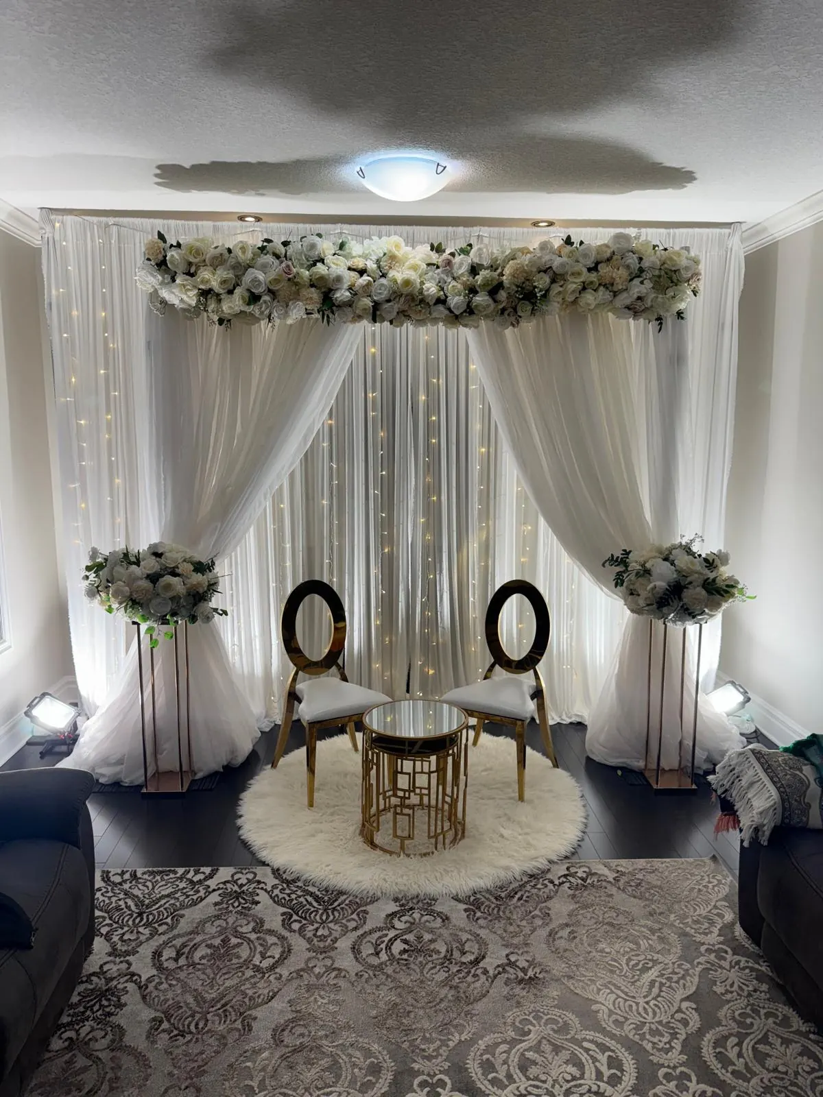 Nikah decor at home: a white draped backdrop with a floral arch, fairy lights, two gold chairs and a gold table on a white rug